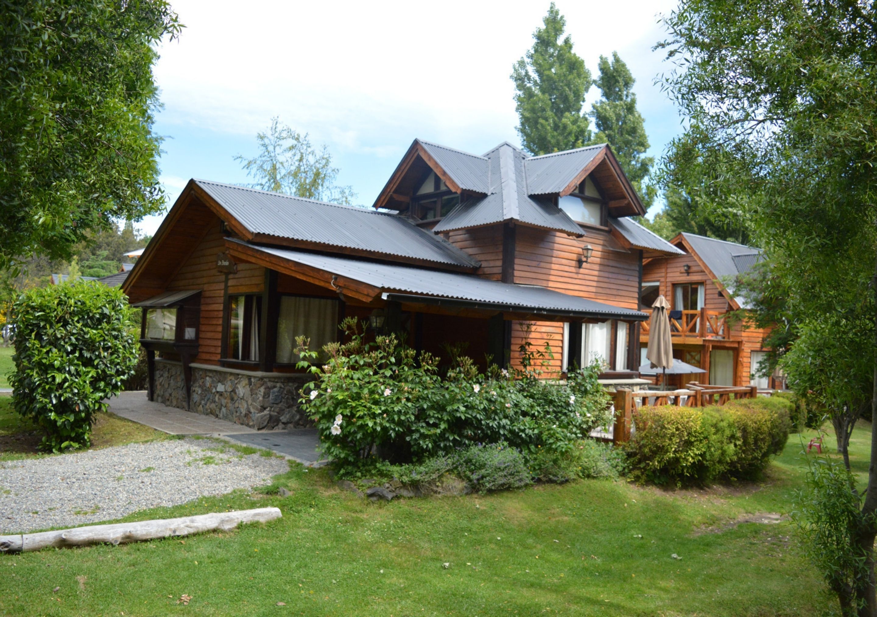 A wooden cabin with a stone foundation, surrounded by lush greenery and a gravel path.