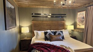 A bedroom with a wooden headboard, two bedside tables with lamps, a bed with a red plaid blanket, and a ski-themed wall decoration. - NEW Pickles Point - Western Mountain Getaway (Grand Lake)