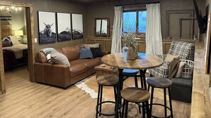 A cozy living room with a brown leather sofa, a round wooden coffee table, and three stools. There are framed pictures on the wall, a window with curtains, and a mirror. - NEW Pickles Point - Western Mountain Getaway (Grand Lake)