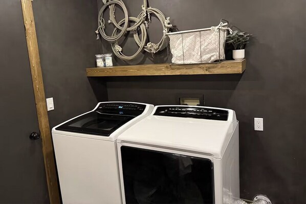A laundry area with two white front-loading washers and dryers, a wooden shelf with a plant and a white bag, and a decorative wall hanging.