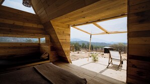 Eine Holzhütte mit geneigtem Dach, einem Bett mit Decke, einem kleinen Tisch und Blick auf eine gebirgige Landschaft.