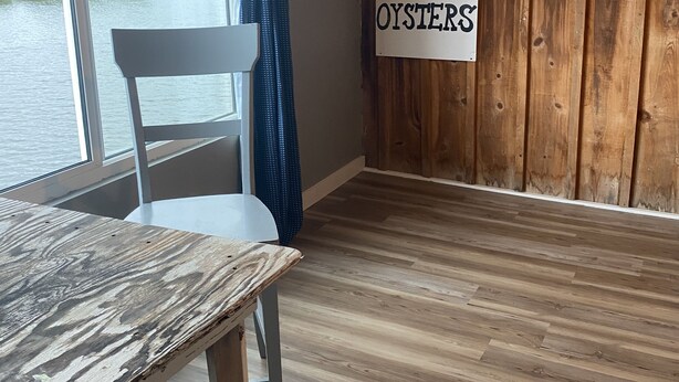 A room with a wooden floor, a white chair, a table, a window with a view of a river, and signs reading "CLAMS" and "OYSTERS". - Charming waterfront 2 bedroom with bonus room that sleeps 8 in scenic Cedar Key (Cedar Key)
