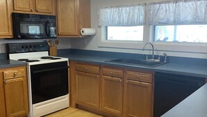A kitchen with wooden cabinets, a microwave, stove, sink, and a window with curtains. - Charming waterfront 2 bedroom with bonus room that sleeps 8 in scenic Cedar Key (Cedar Key)
