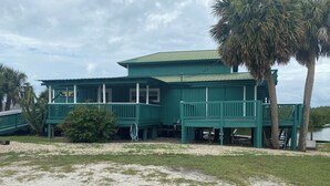 A green building with a balcony, a palm tree, and a body of water in the background. - Charming waterfront 2 bedroom with bonus room that sleeps 8 in scenic Cedar Key (Cedar Key)