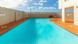 Et rektangulært indendørs swimmingpool med klart, blåt vand, omgivet af en fliseterrasse og en hvid væg med et advarselsskilte.
