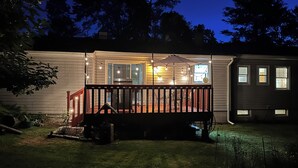 Una casa con terraza de madera, luces colgantes y un espacio cubierto con sillas.
