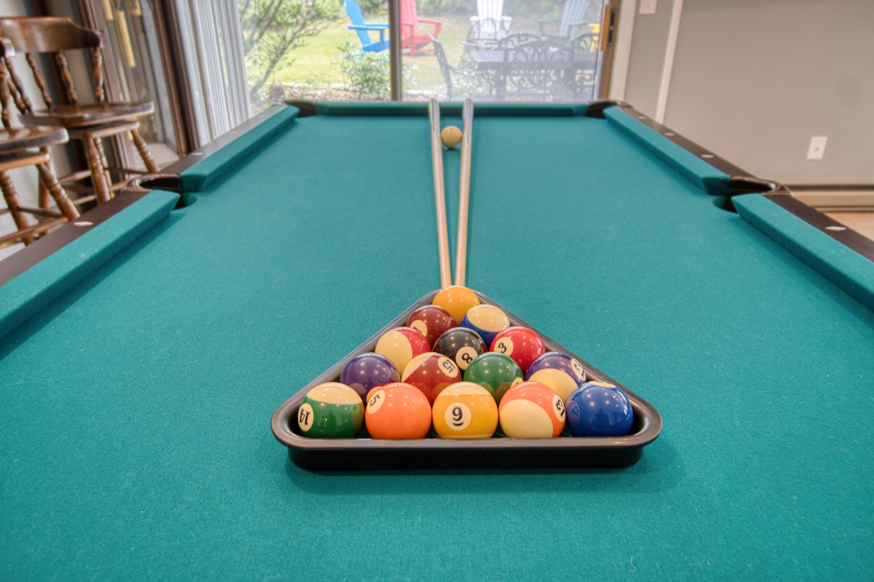 A pool table with a triangular rack of billiard balls, a cue stick, and a window view of outdoor seating.