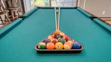 A pool table with a triangular rack of billiard balls, a cue stick, and a window view of outdoor seating.