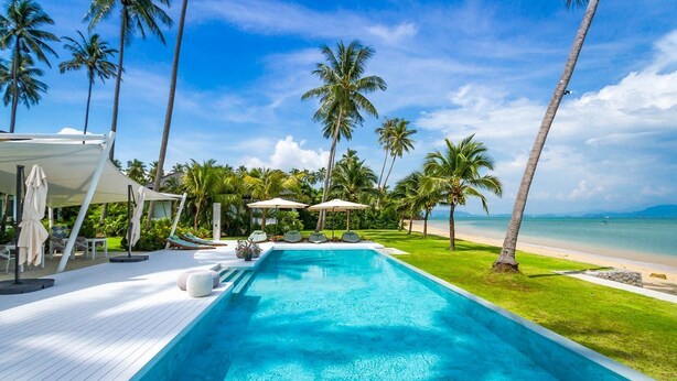 Zona de piscina con sillones de descanso y sombrillas, rodeada de palmeras y vistas a la playa.