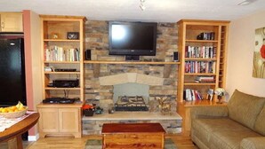 A living room with a stone fireplace, a flat-screen TV mounted above it, wooden bookshelves, a sofa, and a coffee table.