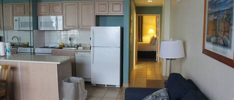 A compact kitchen with a microwave, refrigerator, and stove. There is a trash bin and a blue couch with a pillow.
