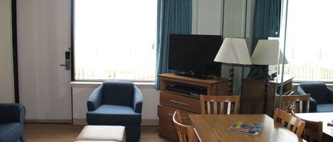 A hotel room with a wooden dining table, two blue armchairs, a sofa, a television, and a lamp.