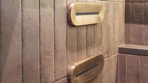 Un panel de control de ascensor metálico con una palanca horizontal y dos botones verticales, frente a una pared con diseño de baldosas verticales.