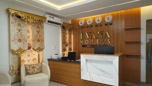 A reception area with a wooden reception desk, a plush armchair, and a wall with clocks and city names. - Evana Hotels (Uyo)