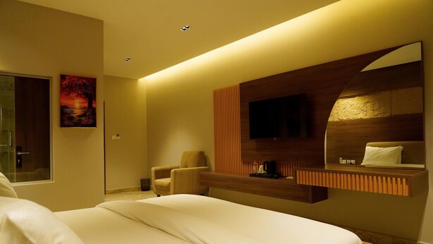 A hotel room with a large bed, a chair, a TV mounted on a wooden panel, and a painting on the wall. - Evana Hotels (Uyo)
