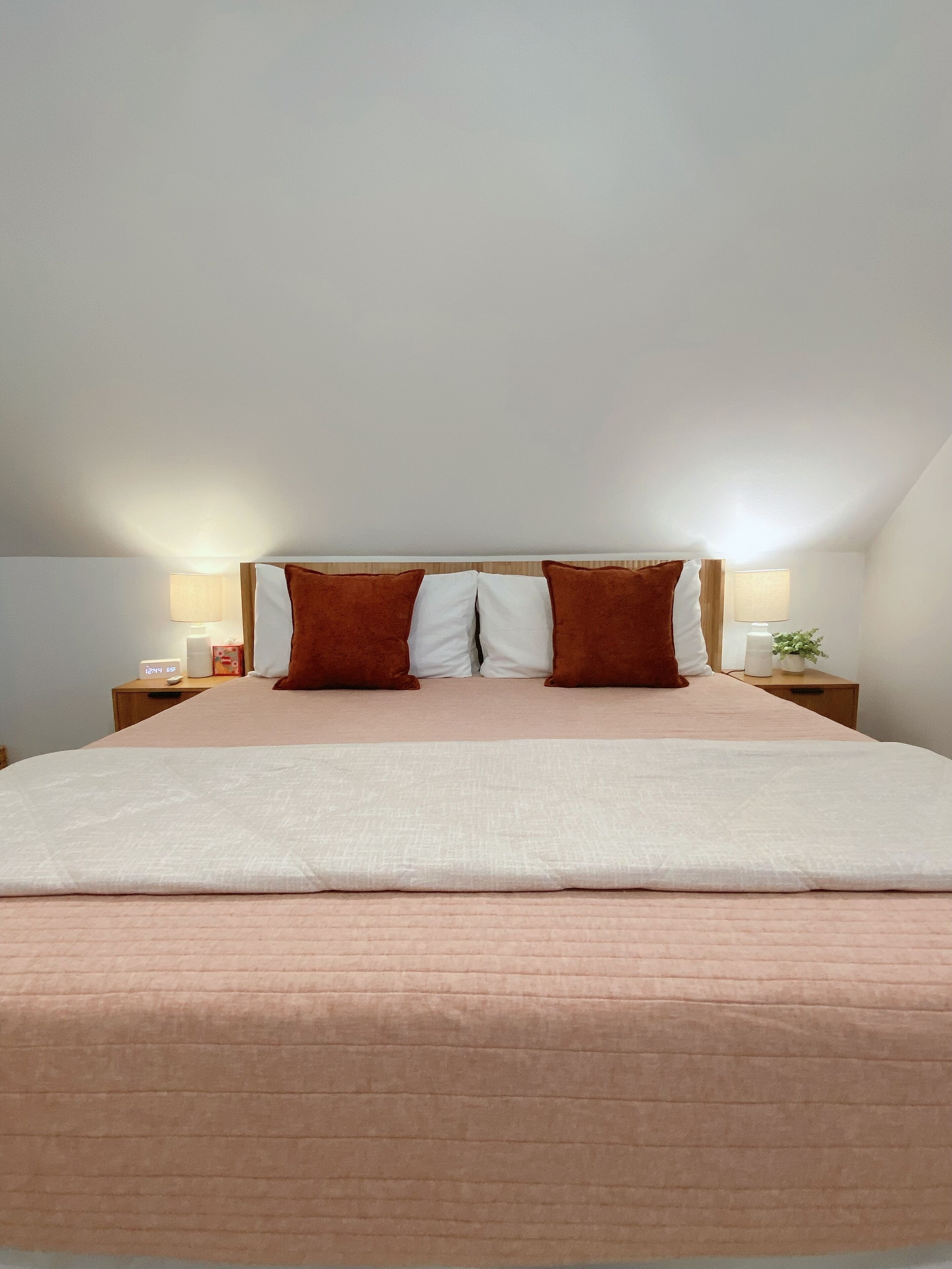 Un lit bien fait, avec une couvre-lit rose et deux oreillers marron, une lampe de chevet et une petite plante posée sur une table de chevet en bois.