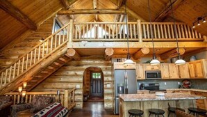 A log cabin interior with a central island kitchen, a loft bedroom, and a staircase leading to the loft.