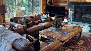 A cozy log cabin interior with a leather sofa, a stone fireplace, and a large window overlooking a forest.