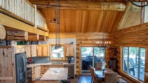 A rustic cabin interior with wooden walls, a kitchen with granite countertops, a dining area with a table and chairs, and large windows offering a view of the outdoors.
