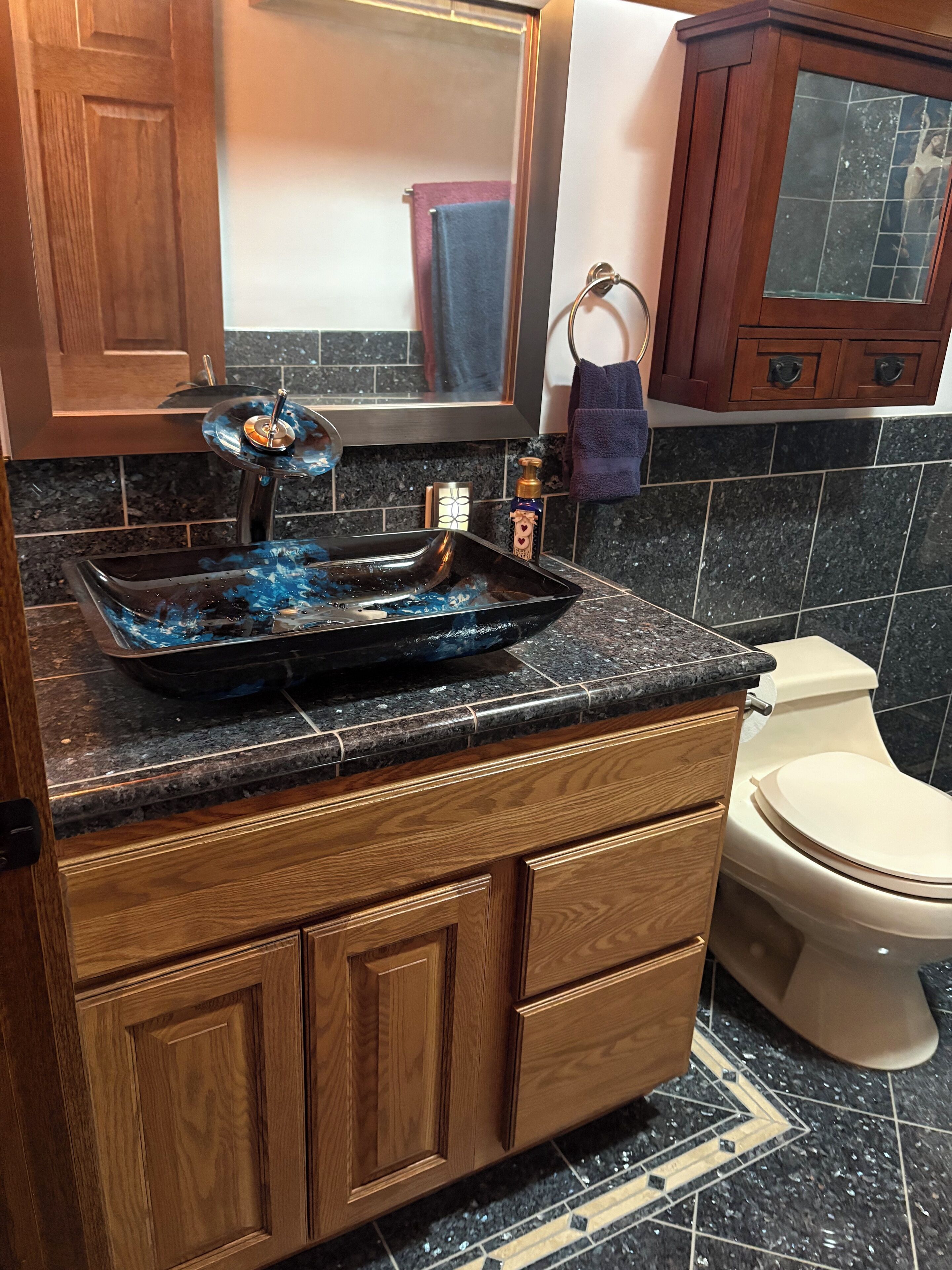 A bathroom with a wooden vanity, a black sink with a blue glass basin, a mirror, and a toilet.