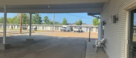 A covered outdoor area with a gravel floor, a white building with a window, and a white truck parked in the background.