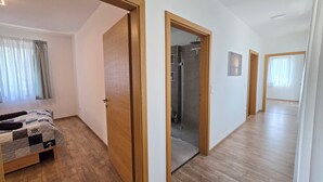 Amplia habitación con piso de madera, una cama con colcha, una puerta que da a un baño con ducha y un pasillo con puerta de madera.