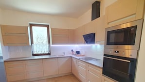 Una cocina moderna con armarios de madera clara, horno empotrado y ventana con vistas a árboles.