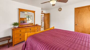 Quarto com cama, cómoda de madeira, ventoinha no teto e uma porta.