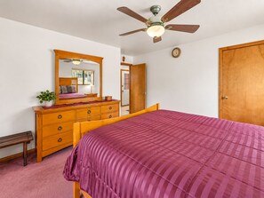 Quarto com cama, cómoda de madeira, ventoinha no teto e uma porta.