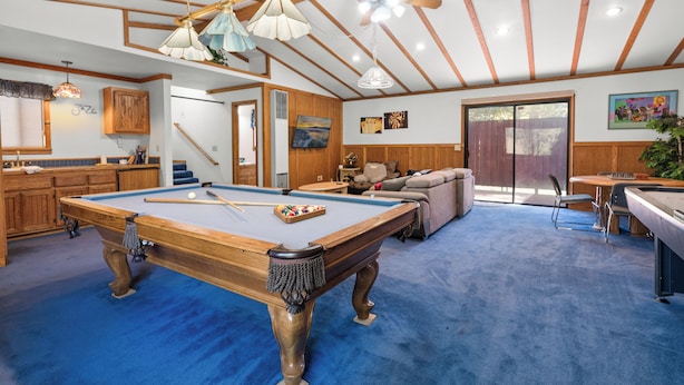A pool table in a room with wooden beams and a blue carpet. There is a sofa and a dining table with chairs. - Silver Rays (Big Bear Lake)