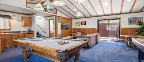 A pool table in a room with wooden beams and a blue carpet. There is a sofa and a dining table with chairs.
