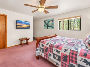 Una camera da letto con un letto, una ventola a soffitto, una finestra vista alberi e due quadri appesi al muro.