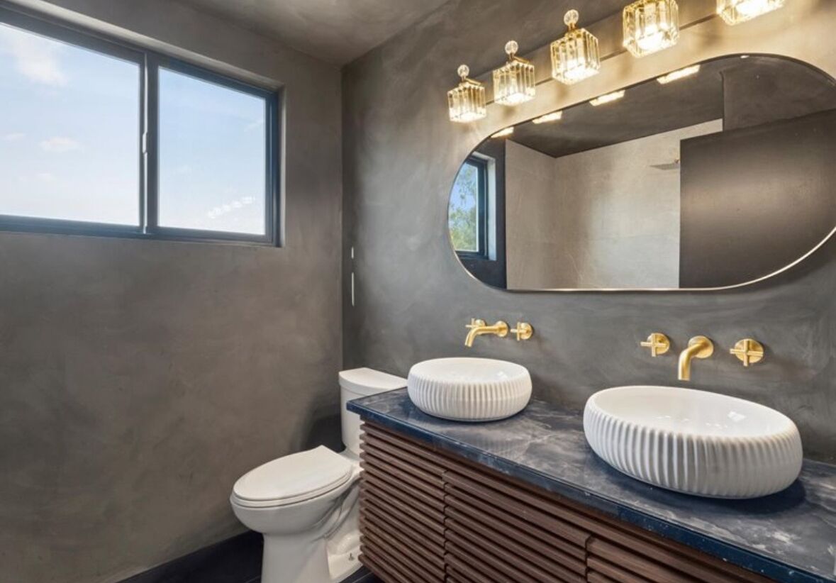 A modern bathroom with a large window, two sinks with gold fixtures, and a vanity with a wooden panel design.