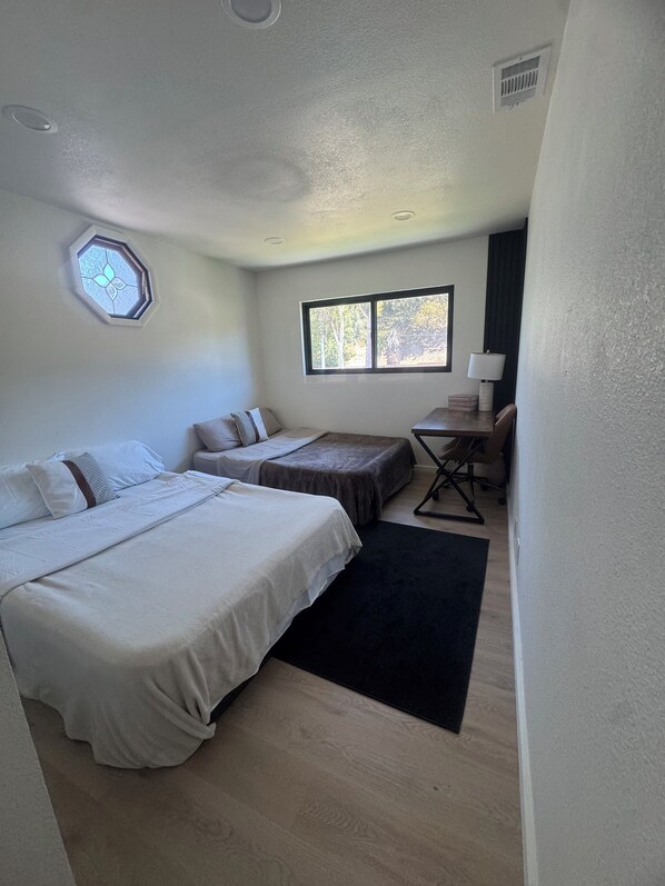 A hotel room with a large bed, a smaller bed, a desk with a chair, a window with a view of trees, and a ceiling vent.