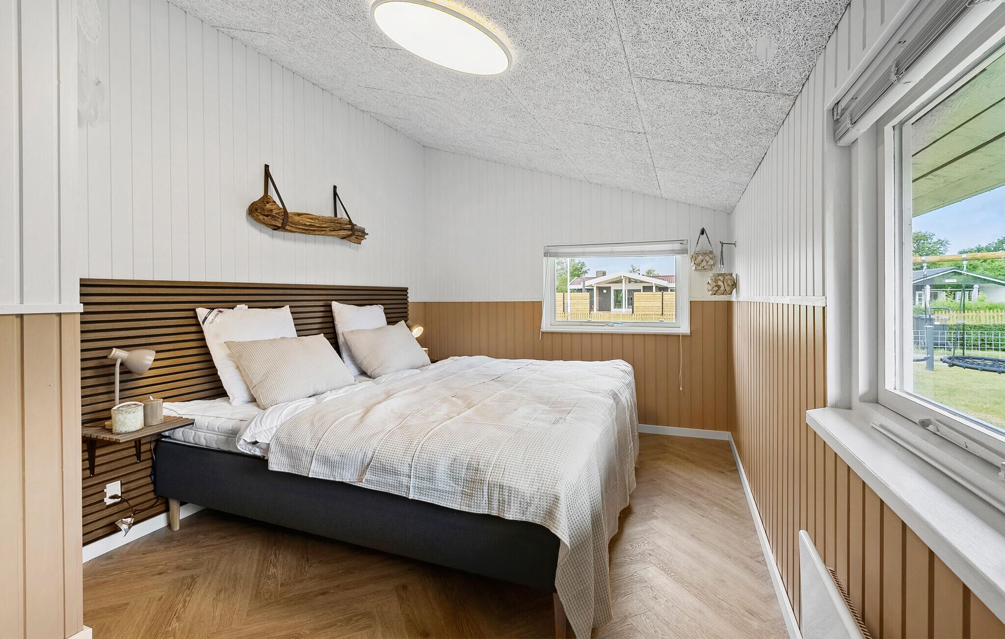 Une chambre à coucher avec une tête de lit en bois, un lit recouvert de linge de lit blanc, une lampe de chevet et une vue sur une maison par la fenêtre.