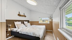 Un dormitorio con cabecera de madera, cama con ropa de cama blanca, lámpara de noche y vista a una casa por la ventana.
