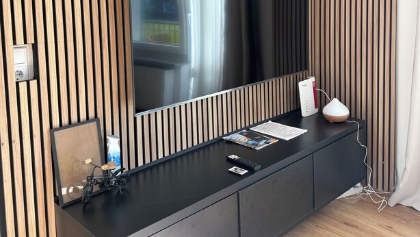 Una sala de estar moderna con un televisor de pantalla plana grande montado en una pared de listones de madera, un mueble de entretenimiento negro con un control remoto, una lámpara blanca y un pequeño objeto decorativo.