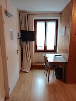 Quarto com piso de madeira, uma mesa de jantar com cadeiras, televisão fixada na parede e uma janela com cortinas.