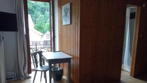 Una habitación con suelo de madera, una mesa con una silla, un cesto de basura pequeño y una cama.