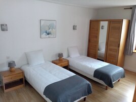 Quarto de hotel com duas camas de solteiro, mesas de cabeceira de madeira, uma pequena mesa, uma luminária na parede, um quadro e um guarda-roupa de madeira.