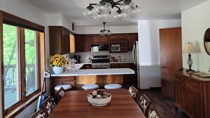 A dining room with a wooden table and chairs, a chandelier, and a kitchen in the background.