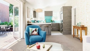 Uma sala de estar moderna com uma poltrona azul, uma mesa de centro de vidro e uma área de cozinha ao fundo.