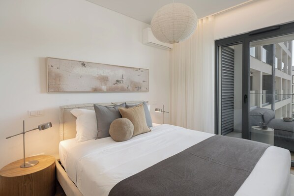 A modern bedroom with a large bed, a bedside table, a wall-mounted artwork, and a view through a glass door.