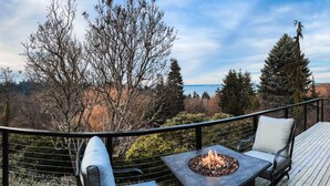 A deck with a fire pit, two chairs, and a view of trees and a body of water. - Vashon View Cottage - Vashon, WA (Vashon)