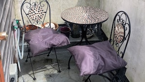A small outdoor patio with a round table and two chairs, a potted plant, and a metal water spigot.