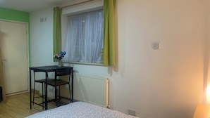 A room with a bed, a desk with a chair, a radiator, a window with curtains, and a door.