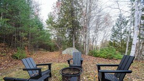 Zona boscosa con tres sillas Adirondack y un hoyo para el fuego.