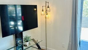 A modern living room with a flat-screen TV on a glass table, a floor lamp, and a window with curtains.