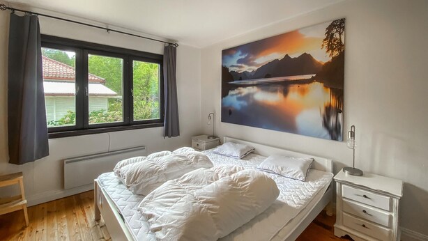 Quarto com cama, mesa de cabeceira, luminária, janela com cortinas e um quadro na parede.