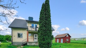 Une maison à deux étages avec un balcon, un grand conifère à l’avant et une grange rouge en arrière-plan.
